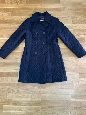 Piretta - Mörkblått , figursydd kappa från Piretta med dubbelknäppning och krage. Kappan har ett quiltat mönster och fickor fram.