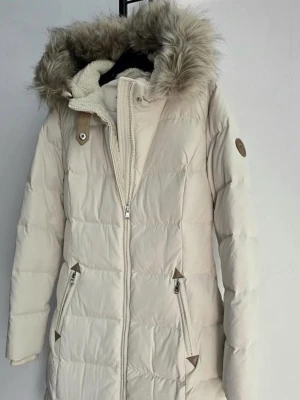 Beige dunjacka från Ralph Lauren - Snygg beige dunjacka från Ralph Lauren med fluffig fuskpäls på huvan och quiltad design. Jackan har dragkedja framtill, två fickor med dragkedja och ett badge på ärmen. Insidan av huvan är fodrad med mjuk teddy. Perfekt för kalla dagar!