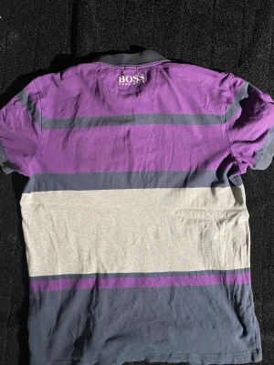 Randig polotopp från Hugo Boss - Säljer en snygg polotopp från Hugo Boss med breda ränder i lila, grått och mörkblått. Toppen har krage, knappslå och logga på bröstet. Perfekt för dig som gillar klassisk stil med en modern twist.