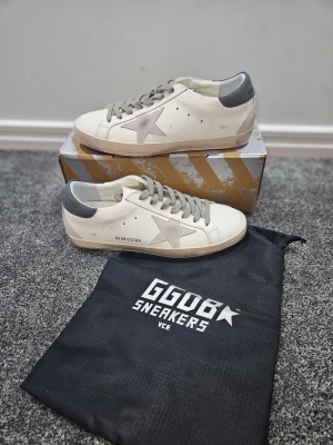 Golden Goose vita sneakers med grå detaljer - Golden Goose sneakers i vitt skinn med grå mockadetaljer och grå snörning. Sidan har en ikonisk stjärna i mocka och hälen är grå med logotyp. Platt sula och rund tå, perfekt för dig som gillar stilrena och coola sneakers.
