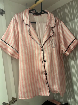 Rosa randig pijamas - Säljer en glansig rosa och vit randig pijamas i storlek XL. Blusen har svarta detaljer vid kanterna, korta ärmar och knappar framtill. På bröstet finns logga i strass. Perfekt för mysiga hemmakvällar. Dock har diamanterna åkt av lite. 