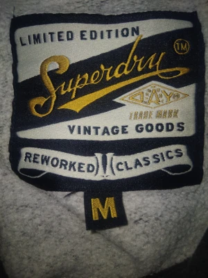 Superdry hoodie limited edition  - Säljer en mörkgrå hoodie från Superdry, Limited Edition Vintage Goods. Stor tryckt logga i orange och vitt på bröstet, med texten 'MOTORCROSS 250 GRAND PRIX'. Klassisk känguruficka och huva. Perfekt för dig som gillar streetwear och snygga detaljer.