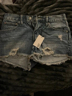 Blå denimshorts från 157 med slitningar - Säljer ett par blå denimshorts från 157 med hög midja och råa, fransiga kanter. Shortsen har slitna detaljer och hål framtill för en cool, avslappnad vibe. Klassisk femficksmodell med knapp och dragkedja. Perfekta för sommarens alla äventyr!
