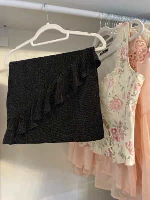 Svart glittrig kortkjol med volang - Svart glittrig kortkjol med snygg volangdetalj diagonalt över framsidan. Perfekt till nyår! Passar XS/S. 