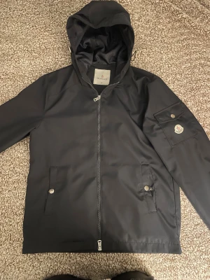 Svart vindjacka från Moncler - Svart vindjacka från Moncler med huva och dragkedja framtill. Jackan har två fickor med tryckknappar och en extra ficka med Moncler-logga på ärmen. Materialet är slätt och syntetiskt, perfekt för blåsiga dagar. Stilren och enkel design som funkar till många outfits. Väldigt bra skick, är ej sönder nånstans.