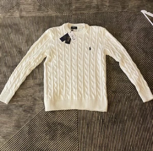 Vit kabelstickad tröja Polo Ralph Lauren - Vit kabelstickad tröja från Polo Ralph Lauren med rund hals och ribbade muddar. Klassisk design med det lilla broderade logot på bröstet. Tröjan har långa ärmar och är gjord i mjuk bomull, perfekt för en clean och stilren look. Helt ny och aldrig använd bara provad men då den sitter liten! Kom dm om frågor och funderingar!
