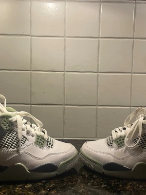 Nike Jordan 4 sneakers vit/grön - Nike Jordan 4 sneakers i vitt och ljusgrönt med svarta detaljer. Skorna har snörning, meshpaneler och en platt sula med synlig Air-enhet. Ovandelen är i skinn och insidan har mjuk textil. Perfekt för dig som gillar klassiska och sportiga sneakers.