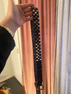 Svart nitad skinnskärp med silverdetaljer - Cool svart Skärp i skinn med massor av silvriga nitar längs hela framsidan. Skärpet har ett rakt snitt och breda axelband, vilket ger en edgy vibe. Perfekt för dig som gillar att sticka ut och vill ha en rockig touch på din outfit.