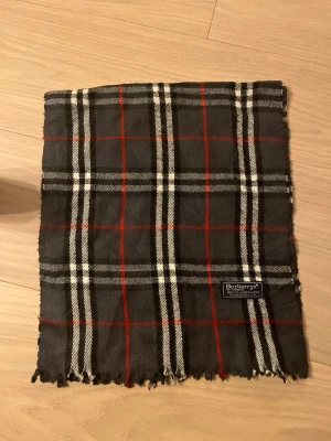 Rutig ullväst från Burberrys - Säljer en klassisk rutig väst från Burberrys i 100% lammull. Västen har en mörkgrå bas med vita och röda rutor, samt fransad nederkant. Märket syns tydligt på etiketten. Perfekt för dig som gillar tidlös brittisk stil.