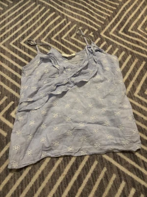 Ljusblå blommig linnetopp med volang - Supersöt ljusblå linnetopp med tunna axelband och broderade vita blommor över hela tyget. Toppen har en v-ringning och en fin volangdetalj framtill som ger extra stil. 