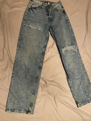  blå jeans med slitningar - Säljer ett par klassiska  jeans i blå med snygga slitningar och hål på benen för en cool vintagekänsla. Hög midja, raka ben och knappgylf. Perfekt för dig som gillar en avslappnad och trendig stil.