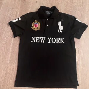 Svart Polo Ralph Lauren piké New York - Svart pikétröja från Polo Ralph Lauren med vit broderad logga, New York-text och stort nummer 5 på ryggen. Guldigt emblem på bröstet och klassisk krage med knappar. Tröjan är i bomull och har korta ärmar. Custom fit-modell som sitter snyggt. Använd 1 gång