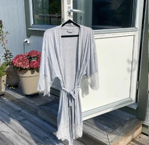 Ljusgrå omlottväst med spetsdetaljer - Säljer en ljusgrå morgonrock med knytband i midjan och snygga spetsdetaljer vid ärmslut och nederkant. morgonrocken är tillverkad i mjukt bomullsmaterial, perfekt för morgonen eller efter duschen. 