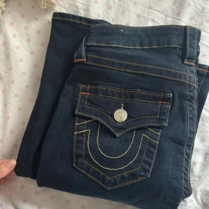 True Religion Becca Bootcut Jeans - Mörkblå jeans från True Religion, modell Becca Mid Rise Bootcut. Snygga kontrastsömmar, klassiska fickor med lock och silverfärgade knappar. Bootcut-ben och mellanmidja ger en tidlös och cool look. Perfekta för dig som gillar detaljer och streetstyle. Helt oanvända köpta för 1299