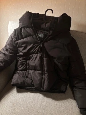 Svart vinterjacka - Svart pufferjacka med stor huva och dragkedja framtill. Jackan har en quiltad design och två sidofickor. Perfekt för kalla dagar! I nytt skick utan prislapp. 