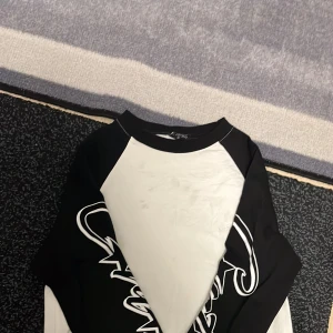 Svartvit långärmad t-shirt med tryck - Säljer en svartvit långärmad t-shirt från Corteiz(r3plica) med coolt grafiskt tryck på ärmarna. Tröjan har kontrastfärgade raglanärmar och rund hals. Materialet känns som mjuk bomull, perfekt för dig som gillar streetwear och vill sticka ut.