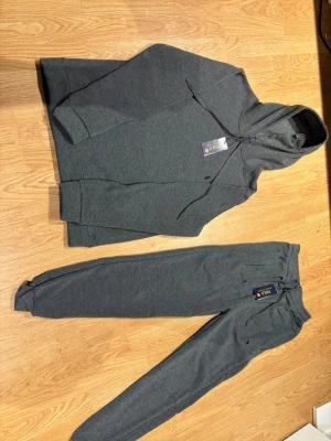 Mörkgrå hoodie och byxorfrån Polo Ralph Lauren - Mörkgrå hoodie från Polo Ralph Lauren med klassisk huva. Broderad svart logga på bröstet och mjukt bomullsmaterial. Perfekt oversized fit och stilren look med ribbade muddar och känguruficka framtill. Byxorna är samma