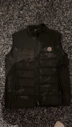 Svart dunväst från Moncler - ⚠️Den ena fickan är sönder invändigt⚠️Säljer en svart dunväst från Moncler med broderad logga på bröstet och stor Moncler-text på ryggen. Västen har två dragkedjefickor fram och quiltad design. Insidan har en cool serietidningsetikett. Perfekt för dig som vill ha en stilren och varm look.