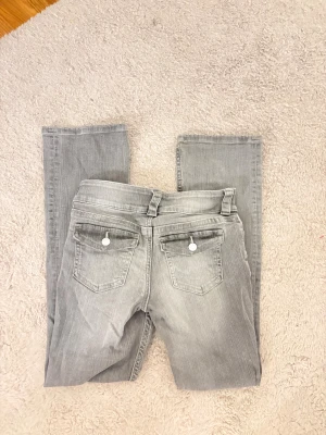 Grå bootcut jeans med silverknappar - Snygga grå jeans med bootcut-snitt och coola silverknappar på bakfickorna. Jeansen har klassisk femficksdesign, bälteshällor och är tillverkade i mjukt jeanstyg. Perfekt för dig som gillar en avslappnad men stilren look.
