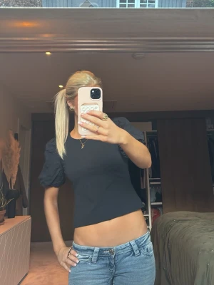 Svart croppad topp med puffärm - Säljer en svart croppad topp med puffiga korta ärmar och rund halsringning. Toppen har en slim passform och är tillverkad i ett mjukt bomullsmaterial. Perfekt att styla med jeans eller kjol för en trendig look.