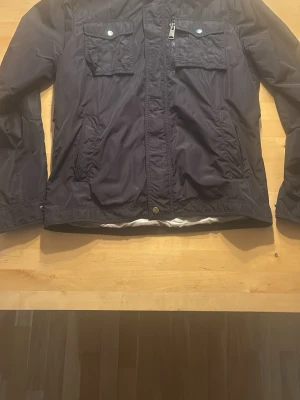 Svart fieldjacka från J.Lindeberg - Snygg svart field från J.Lindeberg med dragkedja och knappar framtill. Jackan har två stora bröstfickor med silverfärgade knappar, samt sidofickor. Jackan har en normal passform med långärmade ärmar.