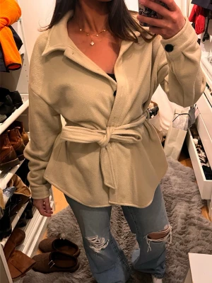 Beige overshirt med bälte och knappar - Snygg beige overshirt med mjukt fleece-material, stora svarta knappar och ett matchande bälte i midjan. Jackan har klassisk krage och långa ärmar med knappdetalj vid mudden. Perfekt att styla med jeans för en chill look