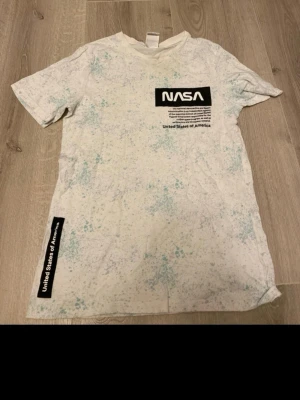 Vit NASA t-shirt från H&M - Säljer en vit t-shirt från H&M med coolt NASA-tryck och texten United States of America. T-shirten har ett stänkmönster i ljusblått och grönt, korta ärmar och rund hals. Materialet är mjuk bomull, perfekt för dig som gillar rymd och streetstyle.