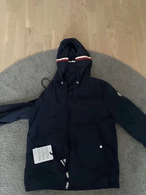 Mörkblå vindjacka från Moncler - Snygg mörkblå vindjacka från Moncler med huva och vita/röda detaljer. Jackan har dragkedja framtill, snörning vid huvan och två fickor med tryckknappar. Moncler-logga på ärmen ger en exklusiv vibe. Perfekt för dig som gillar stilrena och sportiga plagg.