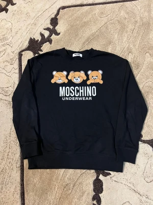 Svart sweatshirt från Moschino -  svart sweatshirt från Moschino med tryck av tre björnar och stor vit logga på bröstet. Tröjan har rund hals, långa ärmar och ribbade muddar. Perfekt för dig som gillar statement-plagg och streetstyle.