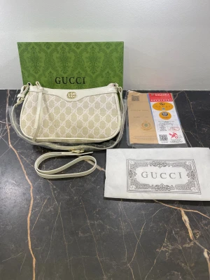 Beige och vit axelväska från Gucci - Snygg beige och vit axelväska från Gucci med klassiskt GG-mönster och guldfärgad logodetalj framtill. Väskan har justerbar axelrem, dragkedja och insida i mjukt material. Perfekt storlek för det viktigaste och riktigt lyxig vibe.