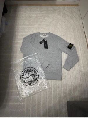 Grå sweatshirt från Stone Island - Snygg grå sweatshirt från Stone Island med klassisk rund hals och ribbade muddar. Tröjan har den ikoniska Stone Island-loggan på vänster ärm och är tillverkad i mjuk bomull. Perfekt för dig som gillar stilrena och trendiga plagg.