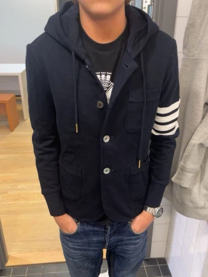 Thom browne hoodie/kavaj - Snygg mörkblå kavaj med hoodie och vita ränder på ena ärmen. Jackan har knappar framtill, snörning vid huvan och flera fickor. Materialet är mjuk bomull och passformen är normal. Perfekt för dig som vill ha en unik och chill stil.