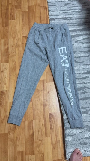 Grå mjukisbyxor från Emporio Armani - Snygga grå mjukisbyxor från Emporio Armani med stort vitt tryck längs benet. Byxorna har elastisk midja med snörning och ribbade muddar vid bensluten. Perfekta för chill eller träning, riktigt sköna och stilrena.