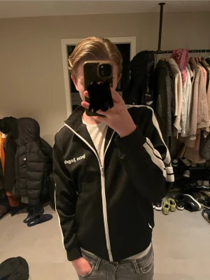 Svart Palm Angels hoodie  - Svart hoodie från Palm Angels med vita ränder längs ärmarna och vit dragkedja framtill. Märkets logga är tryckt på bröstet. Tröjan har en sportig look och är tillverkad i ett mjukt syntetmaterial. Perfekt för dig som gillar streetwear och vill sticka ut. Med normal passform. Ända defekten är dragkedjan som är lite sliten bara, därför påverkar det priset billigare. Annars är tröjan felfri.