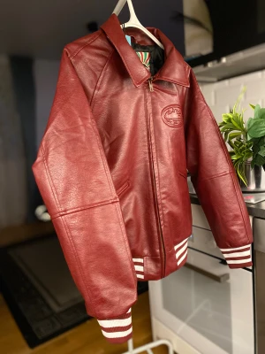 Röd skinn bomberjacka från SkyDive - Säljer en riktigt fet röd bomberjacka i skinn från SkyDive. Jackan har broderad logga på bröstet och ryggen, dragkedja framtill och ribbade muddar med vita ränder. Insidan har coolt mönstrad etikett. Perfekt för dig som vill sticka ut.