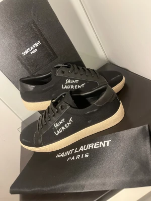  Ysl skor - Säljer ett par svarta Saint Laurent sneakers med vit logotyp på sidan. Skorna har snörning, rund tå och beige platt sula. Ovandelen är i canvas med detaljer i skinn, vilket ger en clean och stilren vibe. Perfekt för dig som gillar exklusiva märken och enkel design.