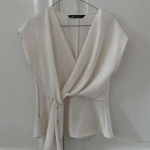 Vit omlottblus från Zara - Snygg vit omlottopp från Zara med knytning i sidan. Toppen har en elegant v-ringning och är ärmlös, vilket gör den perfekt för varmare dagar. Materialet är mjukt och följsamt, vilket ger en skön passform.