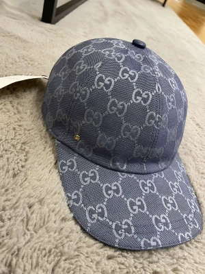 Blå Gucci keps - Snygg blå keps från Gucci med ikoniskt GG-mönster över hela ytan. Kepsen har böjd skärm och justerbar rem baktill i svart läder. Materialet är slitstark bomull och polyester, perfekt för att lyfta din streetstyle. Märkesetikett på insidan.