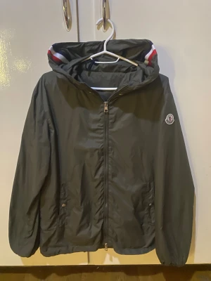 Oliv vindjacka från Moncler - Snygg oliv vindjacka från Moncler med huva och dragkedja framtill. Jackan har Moncler-logga på ärmen och coola röd-vita detaljer på insidan av huvan. Perfekt för dig som gillar stilrena och sportiga plagg med exklusiv känsla.