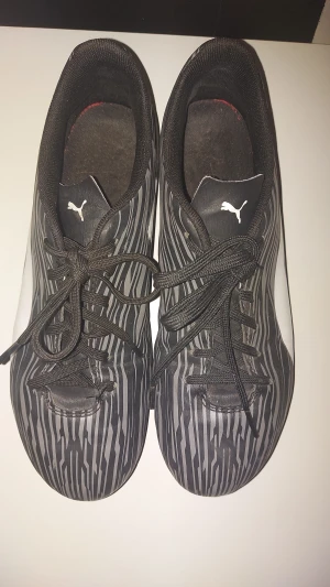 Svarta randiga fotbollsskor från Puma - Säljer ett par svarta fotbollsskor från Puma med gråa och vita ränder och stora vita Puma-loggor på sidan och tungan. Skorna har snörning och platt sula med dobbar för bra grepp på planen. Snyggt mönster och sportig look, perfekta för dig som vill sticka ut på matchen.