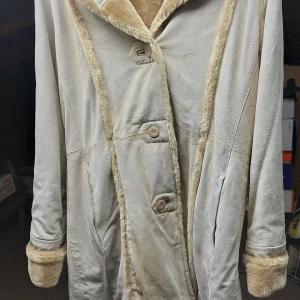 Fuskpälskappa  med fuskpälsdetaljer - Beige fuskpälskappa med mjuka fuskpälsdetaljer längs krage, ärmslut och huva. Jackan har knäppning framtill med stora knappar och en längre, rak passform. 