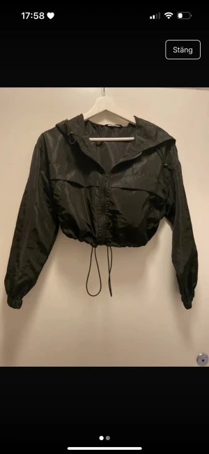 Svart croppad vindjacka med huva - Svart croppad vindjacka med huva och dragkedja framtill. Jackan har snörning i midjan för att justera passformen och elastiska muddar vid ärmsluten. Materialet är glansigt och lätt, perfekt för en chill streetstyle-look.