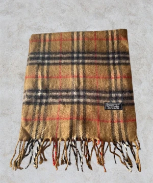 Rutig ullhalsduk från Burberry - Klassisk Burberry-halsduk i 100% lammull. Beige med ikoniskt rutmönster i svart, vitt och rött. Mjuk, varm och i fint skick. Perfekt för både vardag och höst-/vinterstil. Hör av er vid minsta fundering 