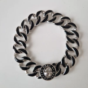 Chunky silverlänk armband Marc Jacobs - Snyggt chunky armband från Marc by Marc Jacobs i silverfärgad metall med svarta detaljer på länkarna. Låset har en rund platta med logotypen ingraverad. Kommer med original svart tygpåse med tryckt logga. Perfekt statement piece för dig som gillar att sticka ut.