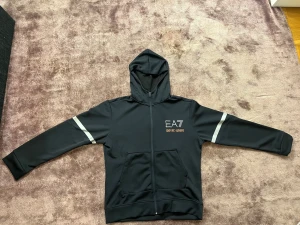 Svart hoodiejacka från Emporio Armani - Svart hoodiejacka från Emporio Armani med EA7-logga broderad på bröstet i vitt och orange. Jackan har huva, dragkedja framtill och två fickor. Snygga vita ränder på ärmarna ger en sportig vibe. Materialet är mjukt och känns som polyester.