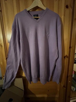 Ljuslila v-ringad tröja Mel Richman XL - Snygg ljuslila v-ringad tröja från Mel Richman i storlek XL. Tröjan har ett broderat emblem på bröstet och är tillverkad i mjuk bomull. Långärmad med ribbade muddar vid ärmslut och nederkant, perfekt för dig som gillar stilrena och klassiska plagg.