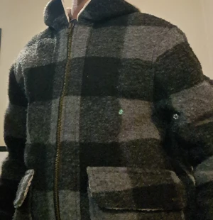 Gråmelerad teddyjacka med Sound of Seattle - Säljer en grå och svart rutig teddyjacka från Trender med broderad text 'SOUND OF SEATTLE' på ryggen. Jackan har dragkedja framtill, stora fickor och mjukt vitt teddyfoder på insidan. Perfekt för dig som gillar streetwear och vill ha något riktigt cozy.