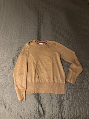 Beige stickad tröja Holly & Whyte - Snygg beige stickad tröja från Holly & Whyte by Lindex i storlek L. Rund hals med kontrastkant i vinrött, ribbade muddar och knappar vid ärmslut. Perfekt för dig som gillar klassisk stil med en twist.