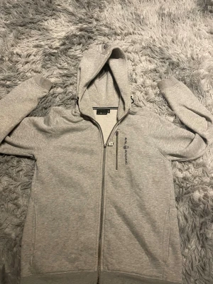 Grå hoodie från Sail Racing med dragkedja - Snygg ljusgrå hoodie från Sail Racing med hel dragkedja och huva. Den har används många gånger och har fläckor på tröjan och ett små hål. Dragkedjan på bröstet är sönder men går att stänga och öppna. Tröjan har en extra zipficka på bröstet och broderad logga i svart. Tillverkad i mjuk bomullsmix, perfekt för chill eller häng. Ribbad mudd vid ärmslut och nederkant för skön passform.