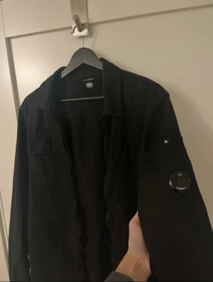Svart overshirt från C.P. Company - Svart overshirt från C.P. Company med klassisk krage, två bröstfickor och dragkedja framtill. Ikonisk linsdetalj på ärmen. Jackan är långärmad och har knappar vid ärmslut samt snörning nedtill. Perfekt för dig som gillar stilrena och funktionella plagg. Den är köpt via vinted och är verifierad, alltså äkta såklart! Skriv vid minsta fundering!